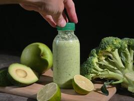 Refreshing green smoothie in bottles on wooden board. Hand picks up smoothie from table with avocado, lime, broccoli, apple, cucumber. Perfect for healthy lifestyle content. Ideal smoothie photo