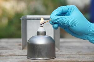 Close up hand wears blue glove, hold a burning match to light fire on alcohol lantern or laboratory lamp. Concept, equipment or tool for science experiment. Educational toolkit in science subject. photo