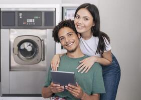 Laundry Service. Happy Young Diverse Couple Posing With Digital Tablet Hugging Near New Washing Machine Indoors, Booking Washer And Smiling To Camera Advertising Laundromat Service photo