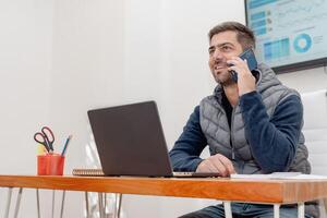 Man talking on phone while working with laptop at his desk. photo
