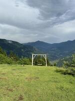 Wooden swing set sits on a grassy hilltop overlooking a stunning mountain range on a cloudy day photo