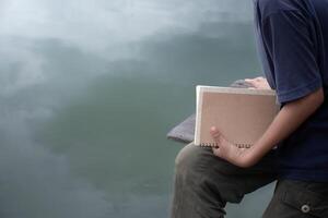 Man holding notebook sitting on wooden bench at lakeside on water surface background,concept of education and work,blank notebook copy space for design,Hand holding blank book,boy holding notebook. photo