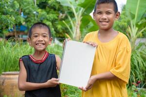 Two boys are smiling charmingly and holding a blank piece of paper or a sign on which to add words or letters in natural forest background at garden for concept of billboard. photo