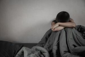A sad boy sits on a black bed in a bedroom giving off a feeling of depression and suffering from domestic violence and childhood bullying. photo