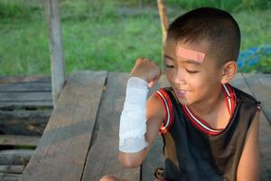 Asian boy smiling shows wounds on his arm and head and bandages them from an accident on a wooden background. Child has accident and wound for first aid concept. photo