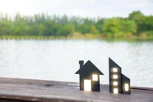 Miniature black house model and commercial building model with dot soft light on wooden planks over green forest and sky background at lake outdoor for concept of home insurance and real estate. photo