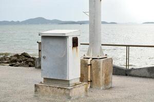 Old rusty electrical control cabinets or white control box and electrical poles sit on concrete floors on the seafront over mountain and sky cloudy background. photo