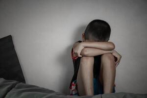 Sad child suffering from depression sitting at bed in bedroom alone due to domestic violence and childhood depression problem in dark tone background. Boy was unhappy and afraid. photo