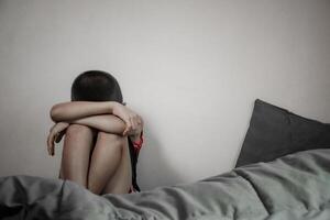 A boy is sitting on his bed with his knees hugged from depression and suffering from domestic violence and bullying in dark tone background. Child sitting alone in a room gives a sad feeling. photo