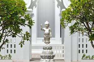 Bangkok Thailand August 7 2023 Baby Buddha Silver Statue Sculpture Clutching Standing on a Lotus Flower in the background a beautiful white architecture Thai temple and green trees. photo