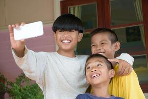 Group of happy and smiling children taking selfies by smartphone camera on background of outdoors. Three boys are holding cell phones to take group photos as a group for activities.