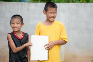 Young asian two boys Wearing a yellow and black shirt was smiling charmingly and holding a white sign or blank white paper to add text on a blurry gray concrete background. photo