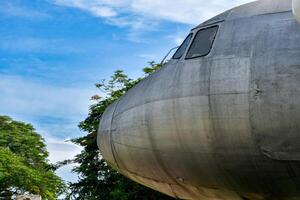 Front view of old airplane head part showing metal surfaces and broken window glass in tree forest and blue sky background. photo