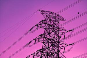 Black high-voltage poles and electric power transmission lines are connected from a power plant on an isolated purple-pink background. photo