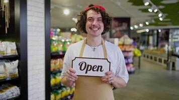 A happy brunette guy with curly hair and a yellow apron holds in his hands a sign with the inscription Open in the supermarket video