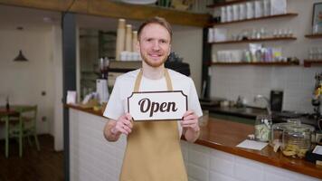 Portrait of a happy brunette guy waiter in a yellow apron who holds a sign in his hands openly in a cafe video