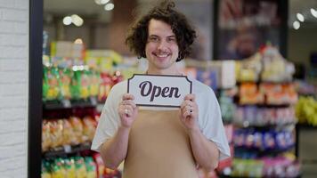 Portrait of a happy brunette guy with curly hair who holds in his hands a sign with the inscription open after the opening of the supermarket video