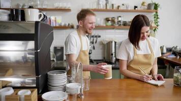 A blond man in a yellow barista apron gives an order to a waitress and they start dancing together at the counter in a cafe video