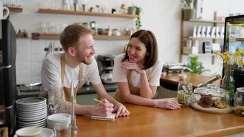 Happy brunette girl in a yellow apron and white T-shirt communicates with her colleague and laughs while writing important thoughts and ideas in a notebook in a cafe video