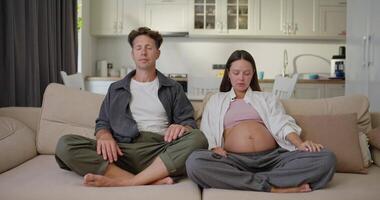 A happy man in a gray shirt together with his pregnant brunette wife sit on the sofa in the lotus position and meditate and then look at each other during their quiet time and waiting for the child at video