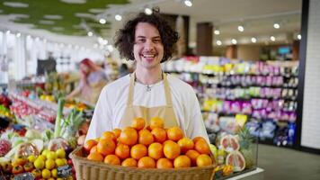 Portrait of a happy brunette guy with curly hair who holds in his hands a box with a lot of orange fruits in a supermarket video