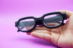 hand holding black frame glasses. black frame glasses isolated on purple background. black frame glasses with curved frames and transparent glass. photo