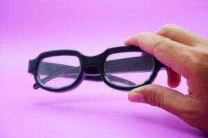 hand holding black frame glasses. black frame glasses isolated on purple background. black frame glasses with curved frames and transparent glass. photo