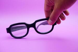 hand holding black frame glasses. black frame glasses isolated on purple background. black frame glasses with curved frames and transparent glass. photo