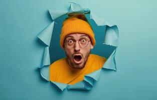 Shocked Man Peering Through Ripped Orange Paper on Blue Background photo