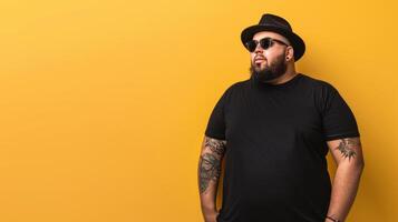A man wearing a black t-shirt and hat stands in front of a bright yellow wall photo