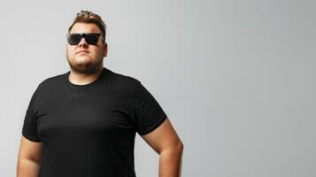 A man in a black t-shirt and sunglasses poses against a plain background photo
