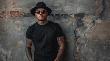 A young man with tattoos and a black fedora leans against a weathered wall photo