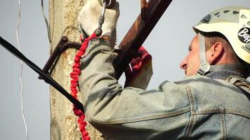 Skilled electrician in helmet fixes wires standing on ladder near high pole against blue sky on summer day backside view. Electrical service and mounting on the pole. Slow motion video