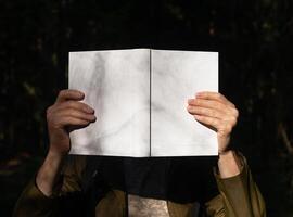. Hand holding white blank open book cover mockup in nature, sunny day. Man hiding head with photo