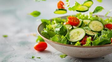 green salad with cucumber and tomatoes falling into a salad bowl against light gray background photo