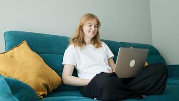 Happy 30s woman making call on laptop while relaxing on a sofa having pleasant conversation waving hand greeting gesture smiling to laptop screen video