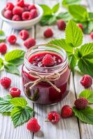 A glass jar of raspberry jam on a white wooden table. Homemade raspberry marmalade and fresh berries and green leaf on the kitchen table photo