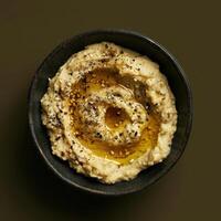 Close up of creamy hummus in black bowl topped with olive oil and sesame seeds. Healthy Middle Eastern food and appetizer concept. photo