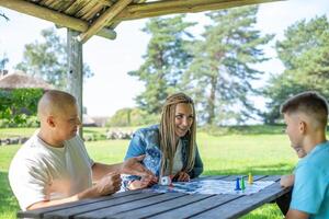 A family is playing a board game on a bright summer day. This scene conveys leisure, strategy, and the quality time spent together, surrounded by lush greenery in a tranquil park setting. photo