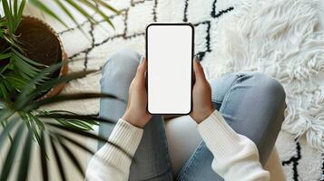 Mockup a person hands holding a smartphone with blank white screen while sitting on the floor at home photo