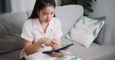 cenas Alto ângulo tiro ,feliz jovem mulher sentado em sofá usando crédito cartão para pagar em digital tábua dentro a vivo quarto às casa ,comprar coisas conectados de tábua, pagar on-line. video