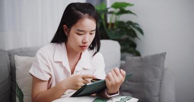 cenas boneca tiro ,feliz jovem mulher sentado em sofá usando crédito cartão para pagar em digital tábua dentro a vivo quarto às casa ,comprar coisas conectados de tábua, pagar on-line. video
