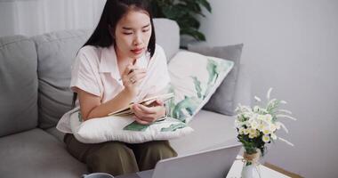 Footage high angle view shot ,Young asian woman explain and take notes while meeting online via laptop on sofa in the living room at home, Working from home video