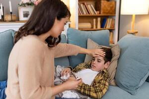 Young woman checking temperature with hand of her ill son. Mother checking temperature of her sick boy. Sick child lying on bed under blanket with woman checking fever on forehead by hand. photo