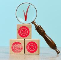 Wooden cubes with red targets stacked on top of each other. On the top cube there is a magnifying glass, through which you can see the red check mark photo