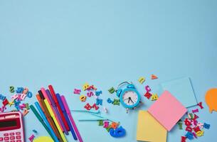 Stationery laid out on a blue background. Multi-colored felt-tip pens, calculator, alarm clock, sticky notes and wooden letters, preparing for study or work photo