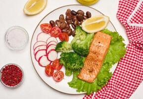 Healthy lunch with grilled salmon on green lettuce, next to vegetables, tomatoes, radishes, broccoli and a portion of beans. photo