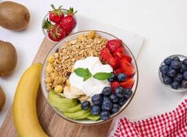 Granola with strawberries, kiwi, banana and blueberries in a round plate on a white table. photo