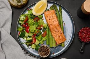 Healthy lunch with grilled salmon with asparagus and fresh tomato and cucumber salad, a row of lemon slice. photo