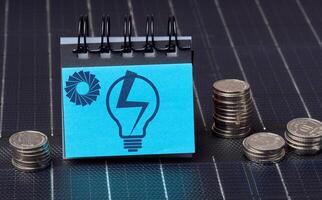 A stack of coins and a notepad with a drawn light bulb stand on the surface of a solar panel, alternative renewable energy concept photo